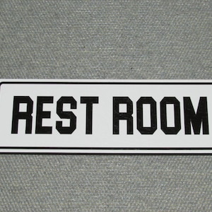 May include: White rectangular sign with black border and black text that reads "REST ROOM".