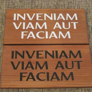 May include: Two wooden signs with the Latin phrase "INVENIAM VIAM AUT FACIAM" written in white and black paint. The phrase translates to "I will find a way or make one."