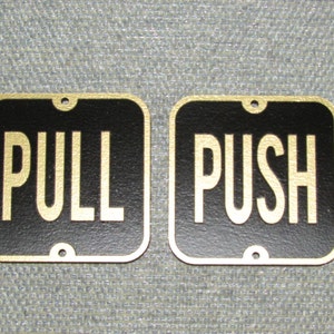 May include: Two black metal signs with gold lettering. The sign on the left says "PULL" and the sign on the right says "PUSH".