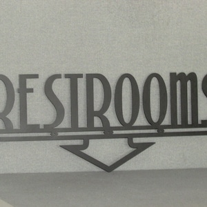 May include: Black metal sign with the word "RESTROOMS" in large, stylized letters. An arrow points down below the word.