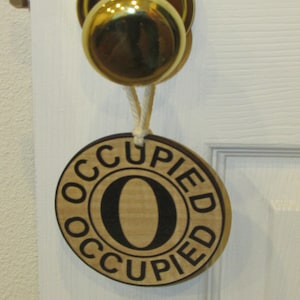 May include: A round wooden sign with the word "Occupied" written in black letters around the edge. The center of the sign has a large letter "O" in black. The sign is hanging from a gold door knob.