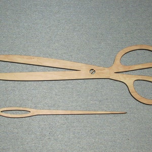 May include: A wooden cutout of a pair of scissors and a needle. The scissors are open and the needle is straight. Both are made of light brown wood.