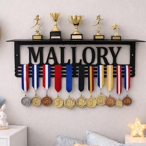 May include: Black metal medal display with the name "MALLORY" cut out. The display holds medals with colorful ribbons and gold trophies. A shelf above holds five gold trophies. The display is mounted on a white wall.