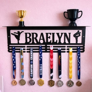May include: Black metal medal display with the name "BRAELYN" cut out. The display has a shelf holding a gold trophy and a black trophy. Below are several medals with colorful ribbons. The wall is pink.