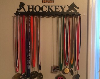 Può includere: Un porta medaglie da hockey in metallo nero con le parole "HOCKEY" e "BOSTON BRUINS" su di esso. Il porta medaglie ha 10 ganci per appendere le medaglie. Ci sono diverse medaglie appese al porta medaglie, tra cui una medaglia d'oro con il numero "13" su di essa.