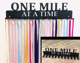 May include: Black metal medal holder with the text "ONE MILE AT A TIME" and hooks for hanging medals. The holder is mounted on a white wall and has several colorful ribbons hanging from it.