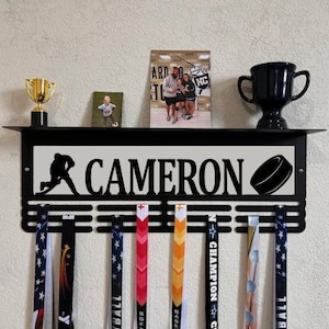 May include: Black metal hockey medal display with the name CAMERON and a hockey puck silhouette. The display has six medal hooks and a shelf for trophies or other items.