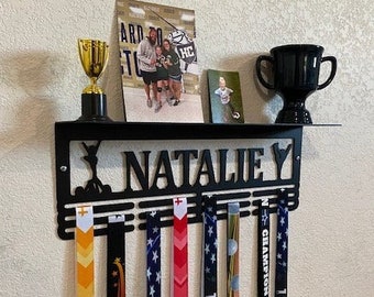 May include: Black metal medal display rack with the name "Natalie" on it. The rack has six hooks for hanging medals. There are five colourful medals hanging on the rack. The medals have the words "Softball", "Gymnastics", "Basketball", "Soccer", and "Cheerleading" on them.