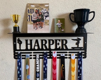 May include: A black metal medal display rack with the name "HARPER" in large letters. The rack has a shelf with two trophies, a photo of a girl, and a photo of three girls. The rack has six medal hangers and is decorated with a gymnast silhouette.