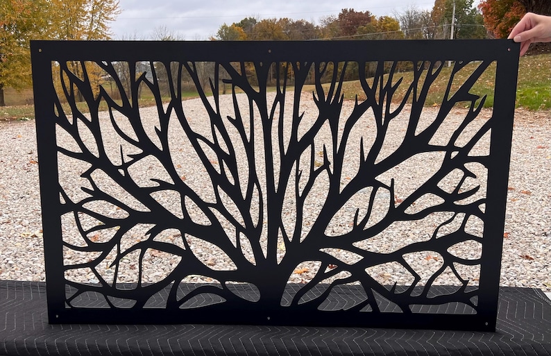May include: A black metal decorative panel featuring a tree branch design. The panel is rectangular and has a cut-out pattern of branches and a tree trunk. The panel is held up by a person's hand.