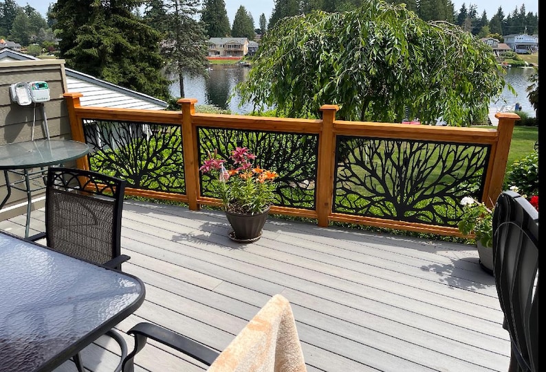 May include: A wooden deck railing with a black metal tree branch design. The railing is made of light brown wood and has a decorative metal panel. A potted plant with colorful flowers sits on the deck. The deck overlooks a lake.