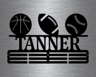 Może przedstawiać: Czarny metalowy stojak na medale sportowe z trzema haczykami do zawieszania medali. Stojak ma wzór koszykówki, piłki nożnej i baseballu. Nazwa "TANNER" jest wyświetlana nad haczykami.