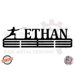 May include: Black metal medal display rack with the name "ETHAN" and a silhouette of a fencer holding a foil. The rack has five horizontal bars for hanging medals. The text "RAW METAL DESIGNS" is below the rack. The words "BEST SELLER" are in a star-shaped seal below the rack. The words "MADE IN THE USA" are in a star-shaped seal below the rack.