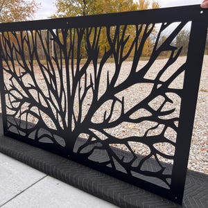 May include: Black metal decorative panel with a tree branch design. The panel is rectangular and features a cut-out pattern of branches and twigs. The panel is held by a hand in the upper right corner, and rests on a black padded surface.