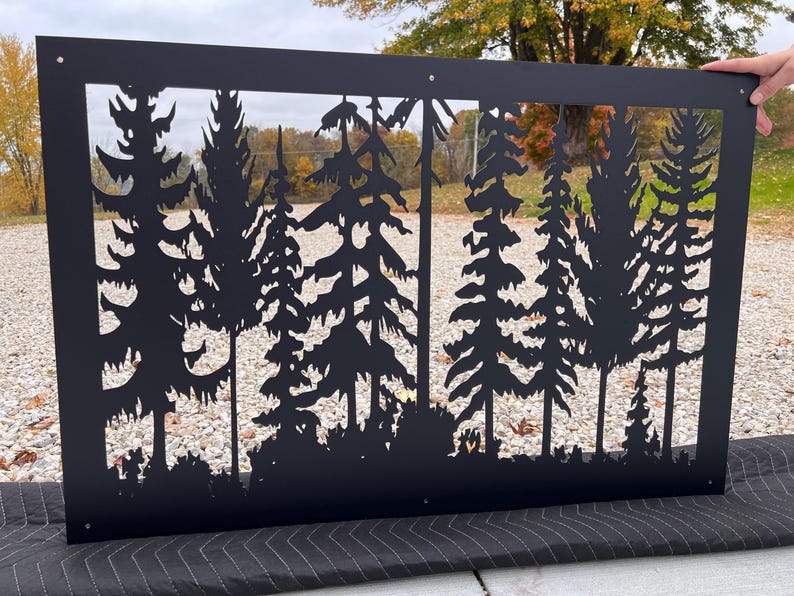 May include: Black metal wall art featuring a forest silhouette design. The rectangular piece has a cut-out of various trees and foliage. The artwork is held by a hand, showcasing the intricate details of the laser-cut design, perfect for outdoor or indoor decor.