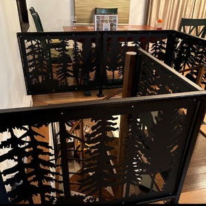 May include: Black metal room divider with a forest silhouette design. The divider is composed of multiple panels, each featuring cut-out images of trees. The panels are framed with a black border and are connected to create a cornered structure.
