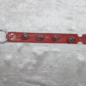 May include: Red leather strap with five silver bells attached. The strap has a floral design and a ring on one end.