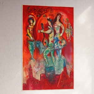 Può includere: Una vivace stampa artistica raffigurante figure colorate con strumenti musicali e un paesaggio urbano. L'opera d'arte presenta uno sfondo rosso e arancione con personaggi stravaganti, una colomba ed edifici. La stampa è montata su una parete bianca.