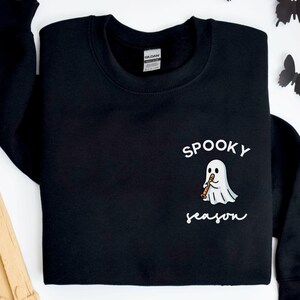 May include: Black crewneck sweatshirt with the words "Spooky Season" and a cartoon ghost playing a musical instrument. The sweatshirt is decorated with black paper butterflies, a skull, and a tombstone.