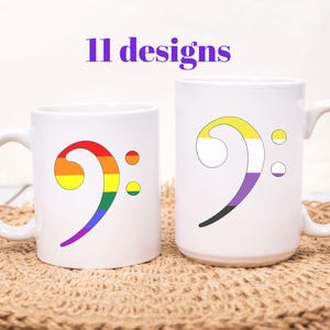 Puede incluir: Dos tazas de cerámica blanca con un diseño de nota musical. Una taza presenta una nota de color arcoíris, y la otra tiene un diseño de bandera del orgullo no binario. El texto "11 designs" está encima de las tazas. Las tazas están sobre una estera tejida.