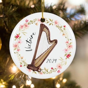 May include: A white ceramic Christmas ornament with a watercolor illustration of a harp and floral wreath. The ornament is personalized with the name "Victoire" and the year "2024".
