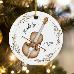 May include: A white ceramic Christmas ornament with a watercolor illustration of a violin and a green wreath with red berries. The ornament is personalized with the name "Madelyn" and the year "2024".