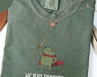 Può includere: Una t-shirt a girocollo color verde muschio con la grafica di una rana verde che indossa un mantello rosso e tiene in mano un trombone dorato. Sotto la rana, il testo recita WE PLAY TROMBONE AT DAWN. L'etichetta Comfort Colors è sul colletto.