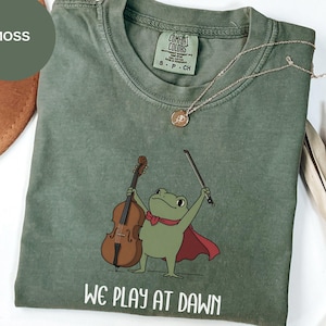 May include: A moss green crewneck t-shirt featuring a graphic of a green frog wearing a red cape and playing a brown double bass with a bow. The text "WE PLAY AT DAWN" is printed in white below the frog. The shirt has a Comfort Colors label.