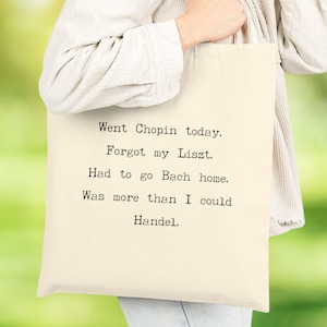 May include: Cream-colored tote bag with black text. The text reads: "Went Chopin today. Forgot my Liszt. Had to go Bach home. Was more than I could Handel." The bag is held by a person.