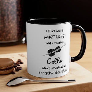 May include: White ceramic coffee mug with a black rim and handle. The mug features a black and white illustration of a cello and the text "I don't make mistakes when playing cello I make spontaneous creative decisions".