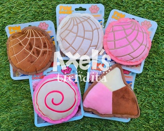 May include: A collection of plush dog toys in the shape of Mexican sweet bread, including conchas and other pastries. The toys are in various colors, such as brown, pink, and white, and are displayed on a green surface.