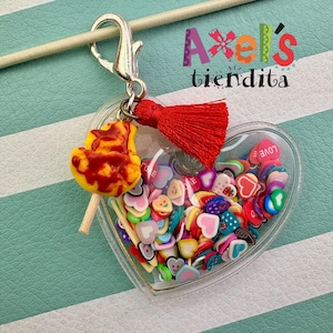 May include: A heart-shaped keychain filled with colorful polymer clay hearts and a lollipop charm. The keychain has a red tassel and a silver clasp. The text "Axel's tiendita" is visible in the background.