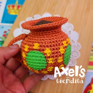 May include: A handmade crochet pot in shades of orange, with yellow and green floral accents. The pot has a small handle and is filled with a dark brown material. The text "Axel's tiendita" is visible in white.