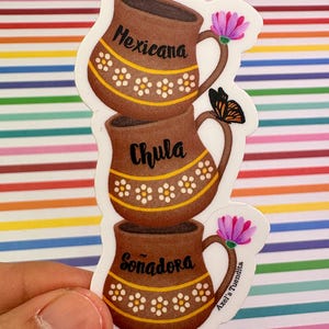 May include: A sticker featuring three stacked brown mugs with the words "Mexicana", "Chula", and "Soñadora". Each mug has a yellow band with white flower details. A pink flower and a butterfly are also present.