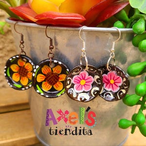 K&ouml;nnte beinhalten: Vier Paar handgefertigte Ohrringe mit floralen Designs. Zwei Paare zeigen orange und gelbe Sonnenblumen, w&auml;hrend die anderen beiden rosa Bl&uuml;ten zeigen. Die Ohrringe h&auml;ngen an einem Metalleimer mit einem farbenfrohen Sukkulentenarrangement.