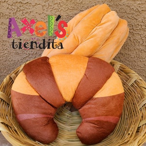 May include: A plush croissant and baguette pillow set in a woven basket. The croissant is brown and orange, while the baguette is a solid orange color. The text "Axel's tiendita" is in the upper left corner.