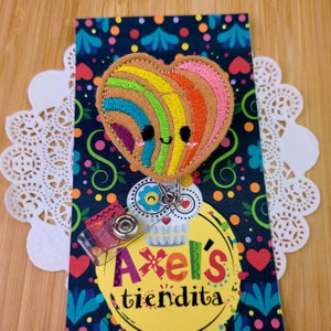 May include: A badge reel in the shape of a heart, featuring a rainbow design and a smiling face. The badge reel is attached to a card with the text "Axel's tiendita" and a colourful floral pattern. The badge reel is made of felt.