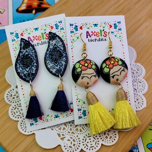 May include: Two pairs of handmade earrings. One pair features blue and white patterned teardrop-shaped pendants with navy tassels. The other pair has round pendants with painted faces, floral headbands, and yellow tassels. The earrings are displayed on cards that read "Axel's tiendita."