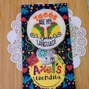 May include: A badge with the text "Tacos are my love language" and two embroidered tacos. The badge is on a colourful card with the text "Axel's tiendita". The background is a wooden surface.