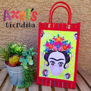 May include: Red tote bag with a Frida Kahlo illustration blowing a bubble. The bag has red handles and trim, with a yellow background. The text "Axel's tiendita" is visible in the upper left corner.