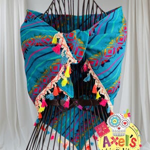 May include: A turquoise textile wrap with colorful geometric patterns and striped accents. The wrap is trimmed with cream-colored fringe and vibrant tassels in shades of pink, yellow, and blue. The wrap is displayed on a black wire mannequin.