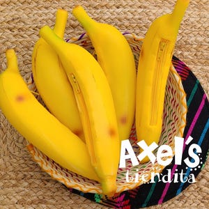 May include: A collection of yellow banana-shaped pouches with zipper closures, nestled in a woven basket. The bananas have brown spots and are arranged on a woven surface. The text "Axel's tiendita" is visible.