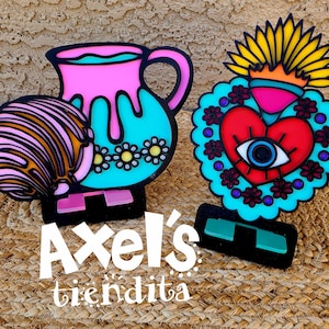 May include: Two colorful acrylic cutouts. One is a pink and turquoise pitcher with a black outline and flower details. The other is a red heart with an eye, surrounded by a turquoise border and a yellow crown. Both are on black bases.