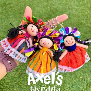 May include: Three handmade doll keychains with colorful ribbon hair and traditional dresses. Each doll has a smiling face and braided black hair. The dolls are attached to silver keyrings. The text "Axel's tiendita" is at the bottom.