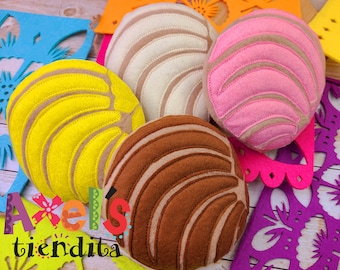 May include: A collection of colorful, felt concha bread pillows in yellow, pink, brown, and cream colors. The pillows have stitched details and are arranged with decorative paper cutouts. The text "Axel's tiendita" is visible.