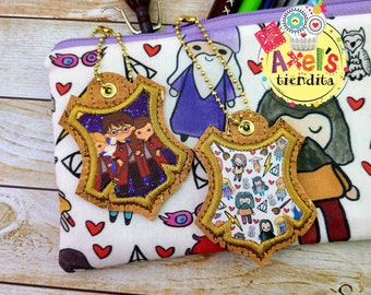 Op de afbeelding: Twee schildvormige kurken sleutelhangers met gouden balletjeskettingen. De ene toont een trio stripfiguren in gewaden, de andere een patroon van kleine magische iconen. Ze liggen op een wit etui met een print van stripfiguren en hartjes.