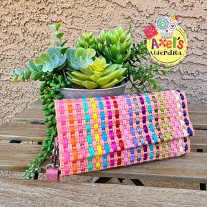 May include: A rectangular clutch bag made of woven plastic strips in a repeating pattern of light pink, orange, turquoise, yellow, red, purple, and blue. The bag features a silver metal clasp and a detachable pink wrist strap on the side.