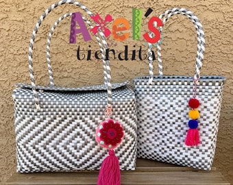 May include: Two woven tote bags with gray and white diamond patterns. One bag has a flap closure and a pink tassel with a flower charm. The other bag has a pink, yellow, and blue pom-pom tassel. Both have braided handles.