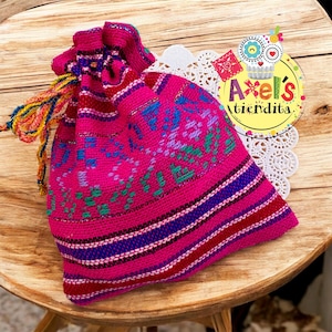 May include: A vibrant pink drawstring bag with intricate woven patterns in blue, green, and red. The bag features a colorful tassel and the text "Axel's Tiendita" in a circular logo. The bag is placed on a wooden surface.