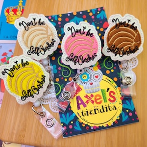 May include: Badge reels embroidered with concha designs in white, yellow, pink, and brown, each bearing the text "Don't be Self Conchas." The reels are attached to clear plastic holders. A colorful card with the text "Axel's tiendita" is also visible.